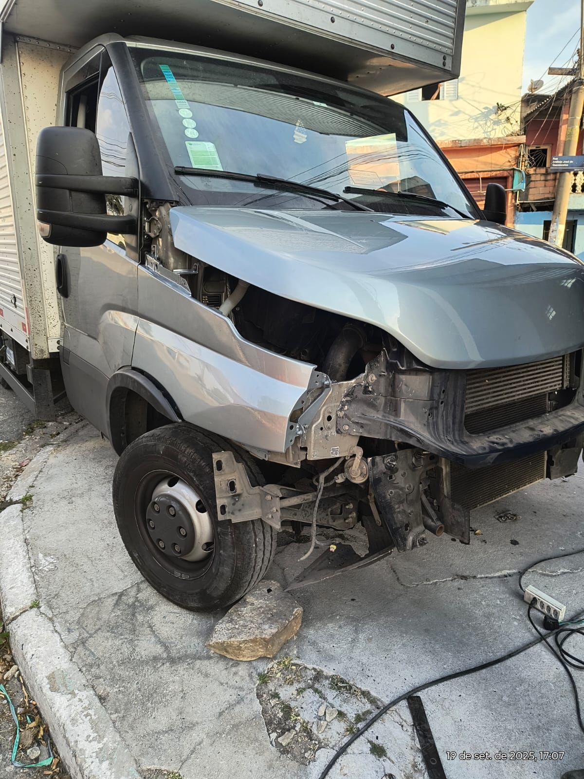 caminhão Iveco Daily prata na rua com o para-choque e a frente parcialmente desmontados, revelando o motor e o radiador